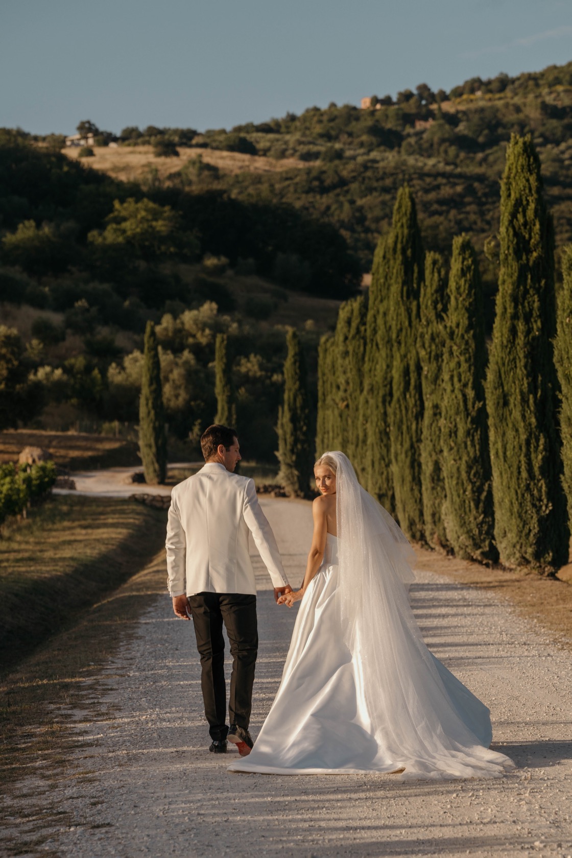 Fern and James walking hand in hand along the cypress-lined gravel drive at golden hour, rolling hills behind them