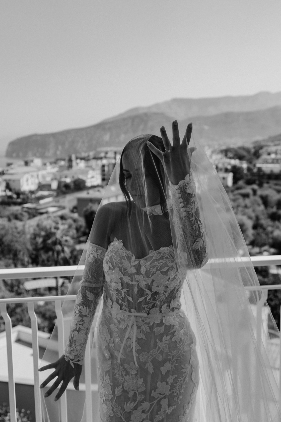 Black and white bridal portrait of Jade in a long-sleeve floral lace gown and cathedral veil on a Sorrento balcony, coastline and hills visible behind her