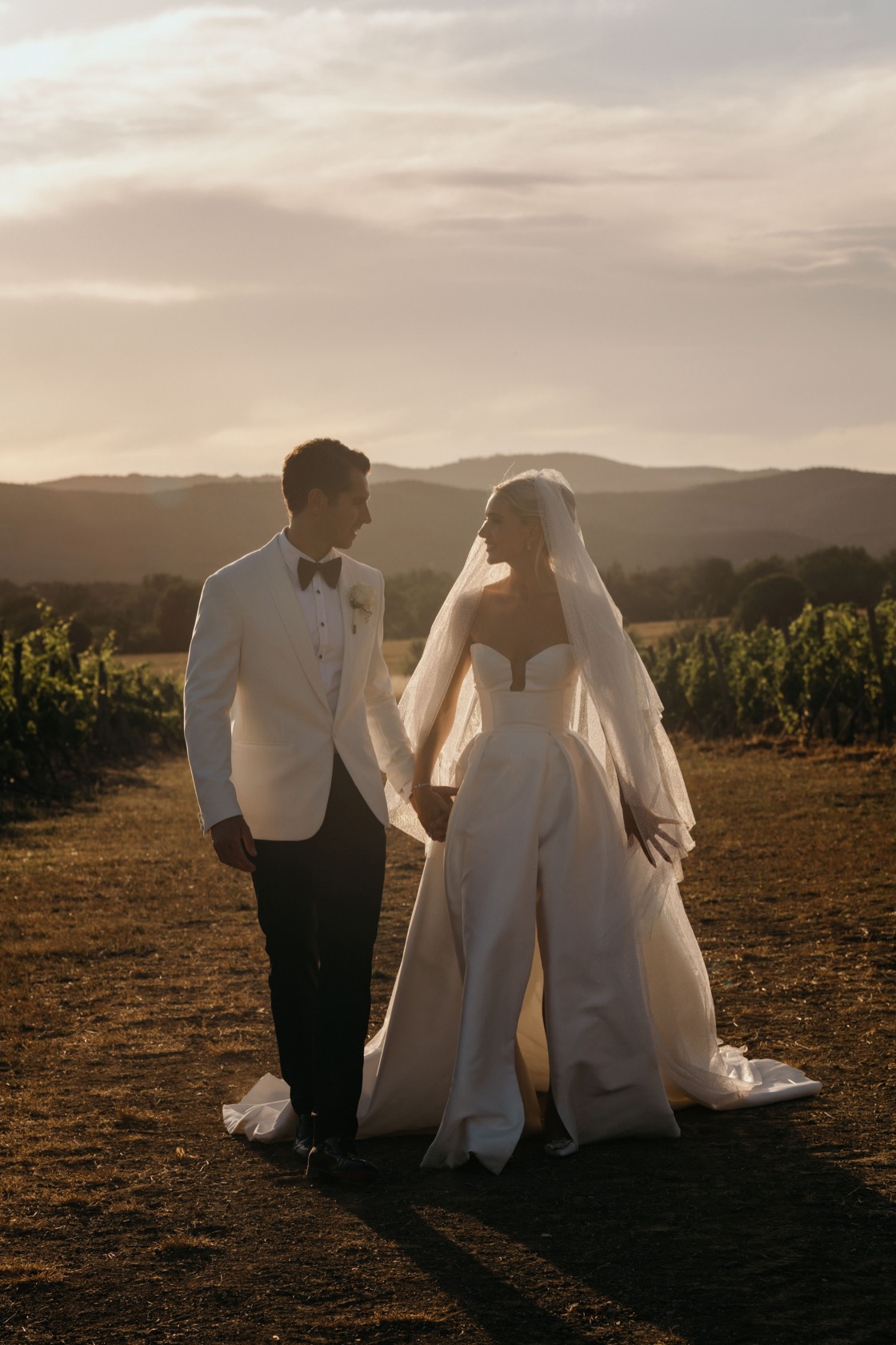 Fern and James standing among vineyard rows at sunset, rolling Tuscan hills behind them at Conti di San Bonifacio Wine Resort