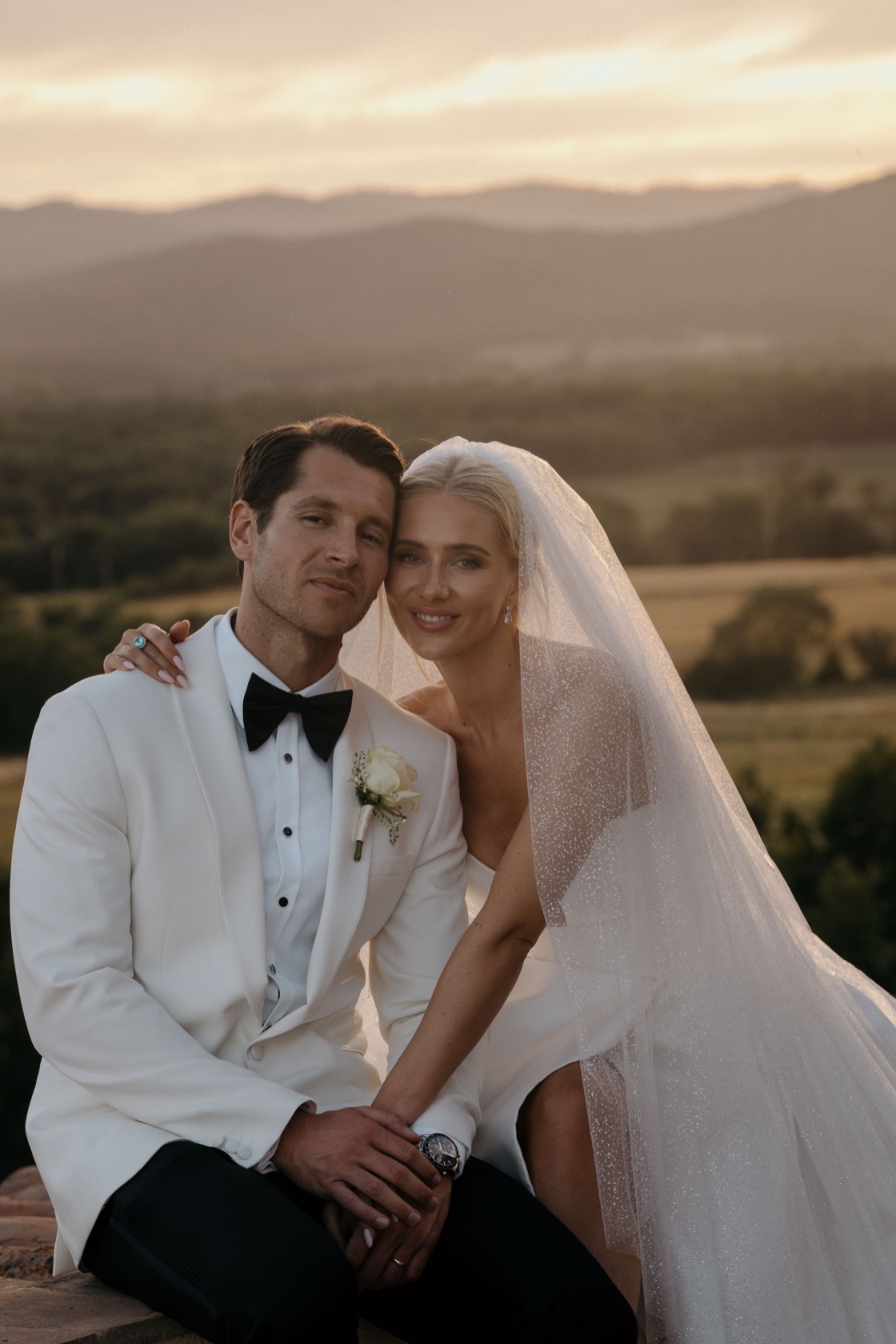 Fern leaning her head on James's shoulder on a stone ledge, glitter veil, rolling Tuscan hills and warm sunset sky behind them