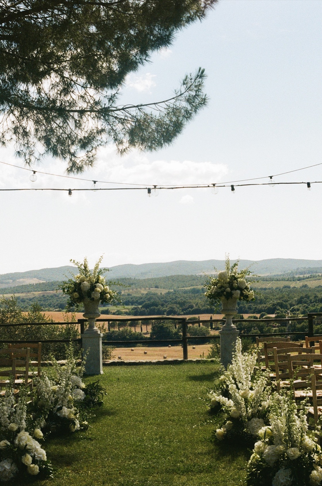 Outdoor ceremony setup at Conti di San Bonifacio, white florals lining the grass aisle, stone urns, festoon lights and Maremma hills beyond