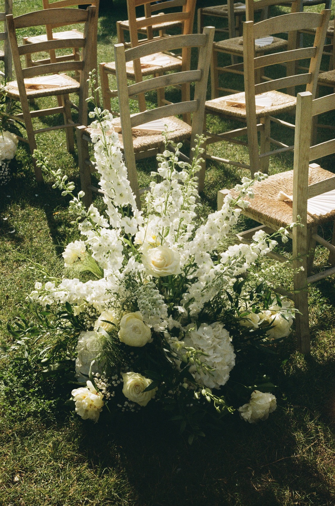 Large white floral arrangement beside rattan ceremony chairs, featuring white delphiniums, garden roses, hydrangea and ranunculus