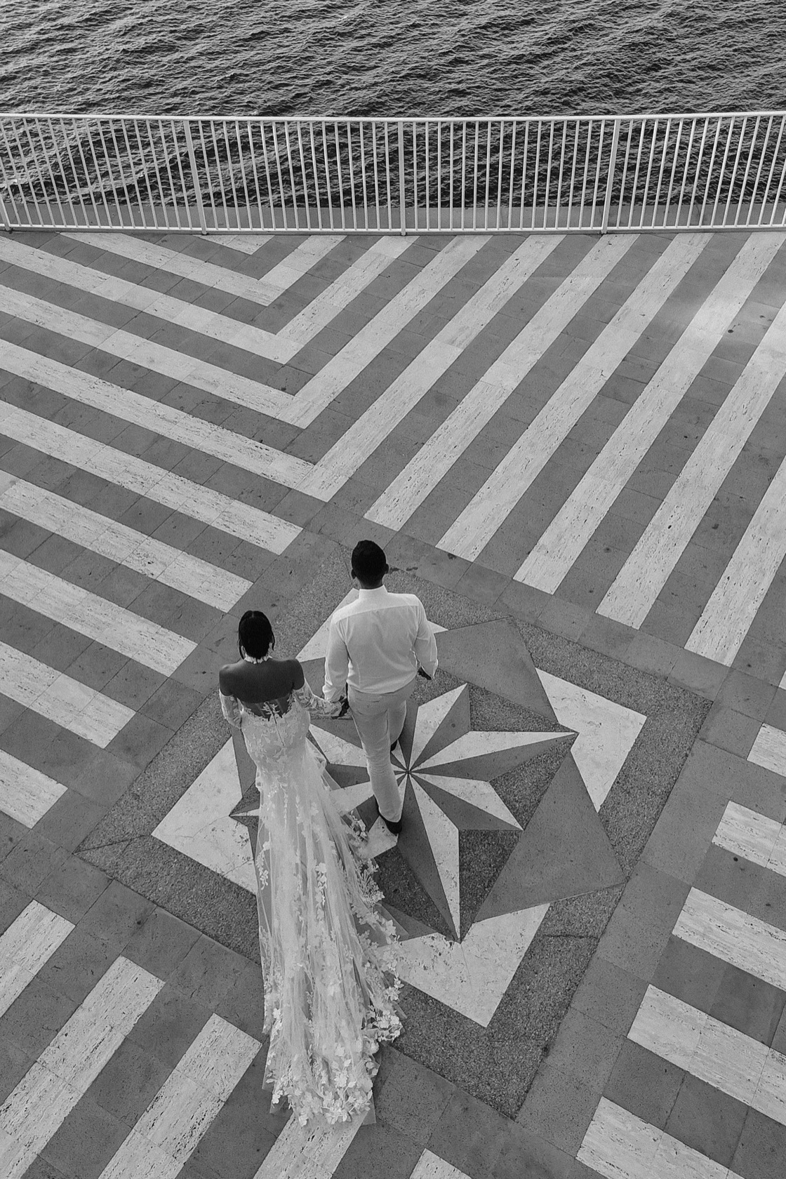 Aerial black and white photograph of Jade and Matt hand in hand on the geometric patterned marble terrace at The Cloisters, Sorrento, overlooking the sea — long-sleeved lace gown and cathedral train