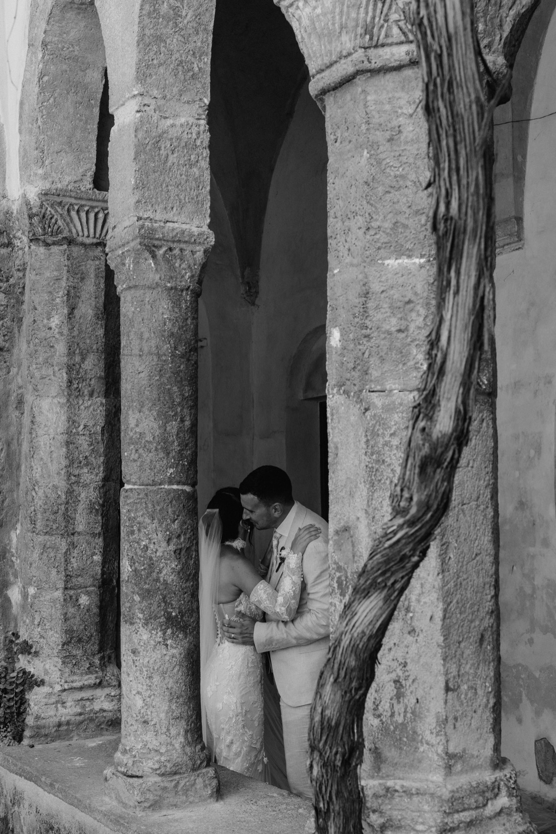 Jade and Matt embracing and kissing in a shadowed archway between ancient stone columns at The Cloisters — twisted tree trunk in the foreground, black and white fine art portrait