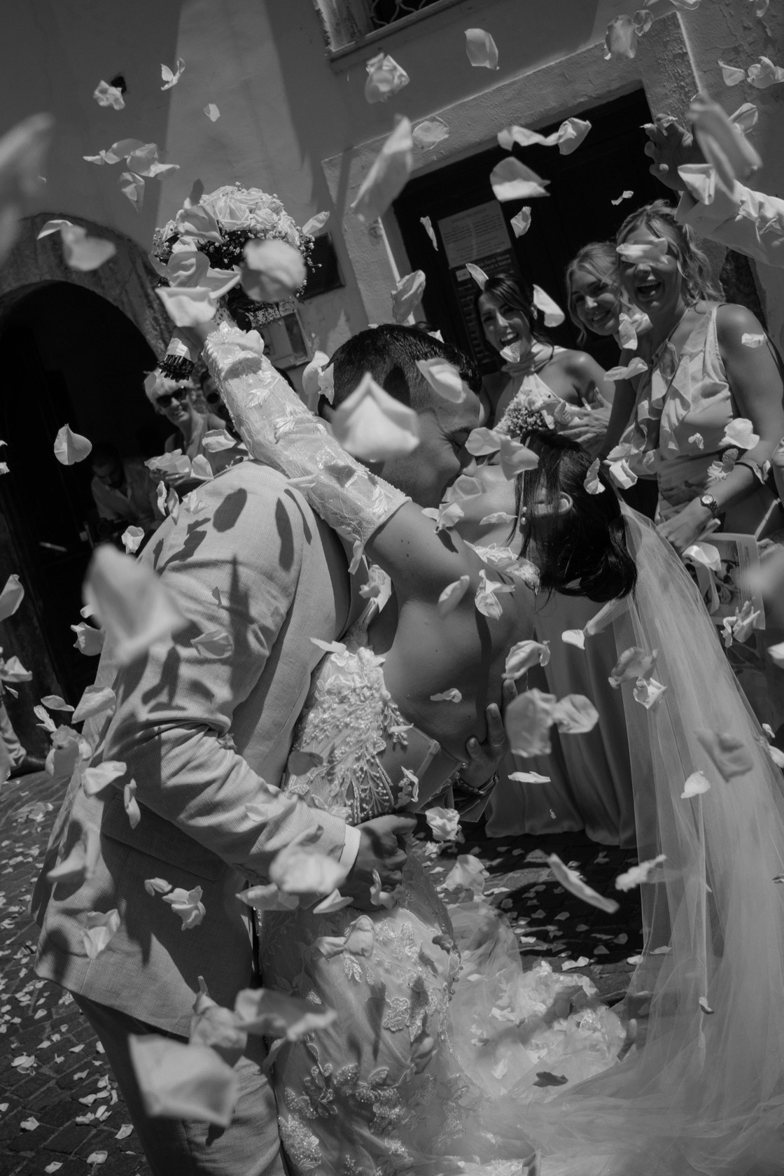 Bride and groom share their first kiss in a shower of rose petals thrown by bridesmaids outside the stone doorway of The Cloisters, Sorrento, Italy.